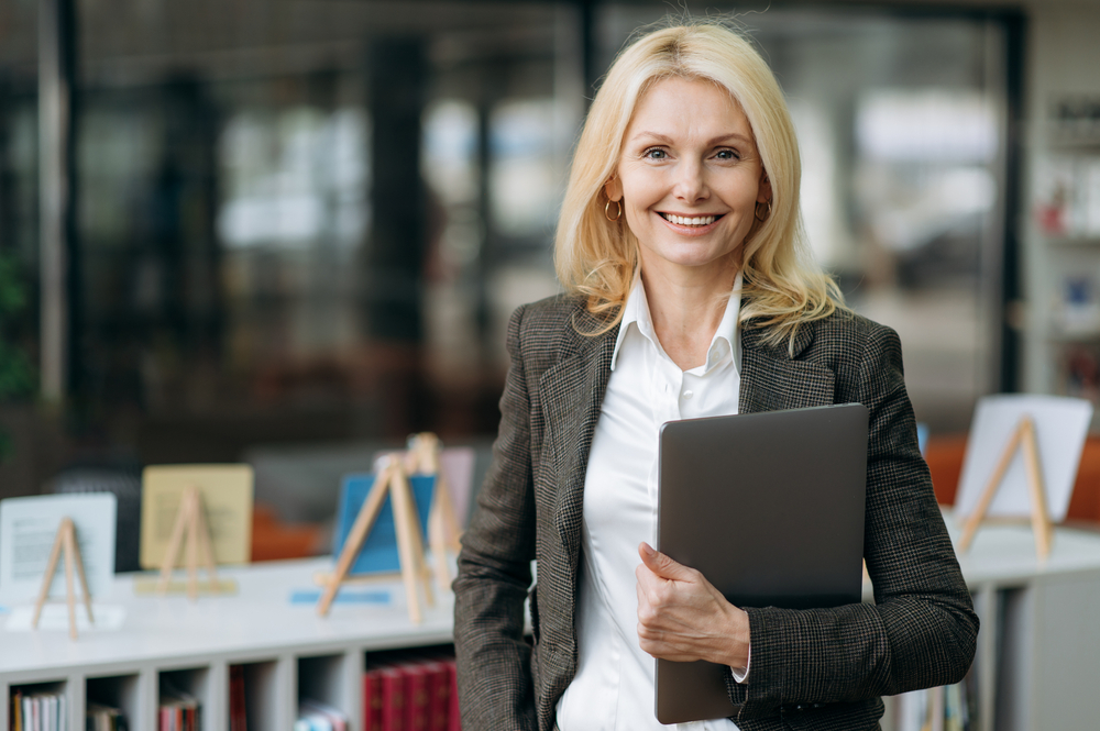 Professional woman in a business suit holding a laptop, smiling confidently in an office setting.