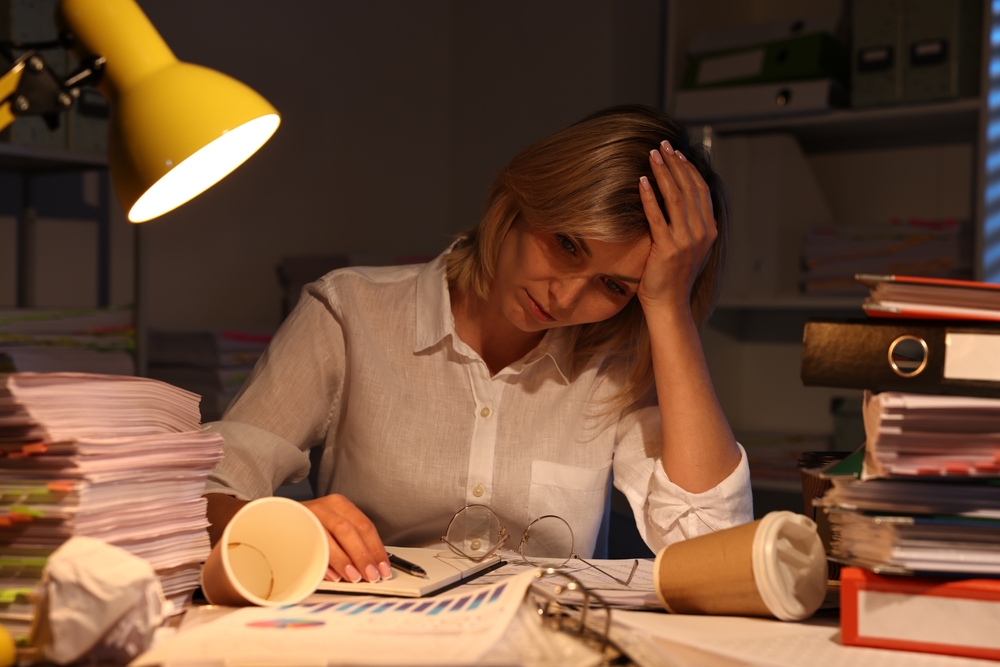 Australian professional woman working late at her desk, surrounded by files and paperwork, showing the stress and self-doubt associated with the impostor phenomenon